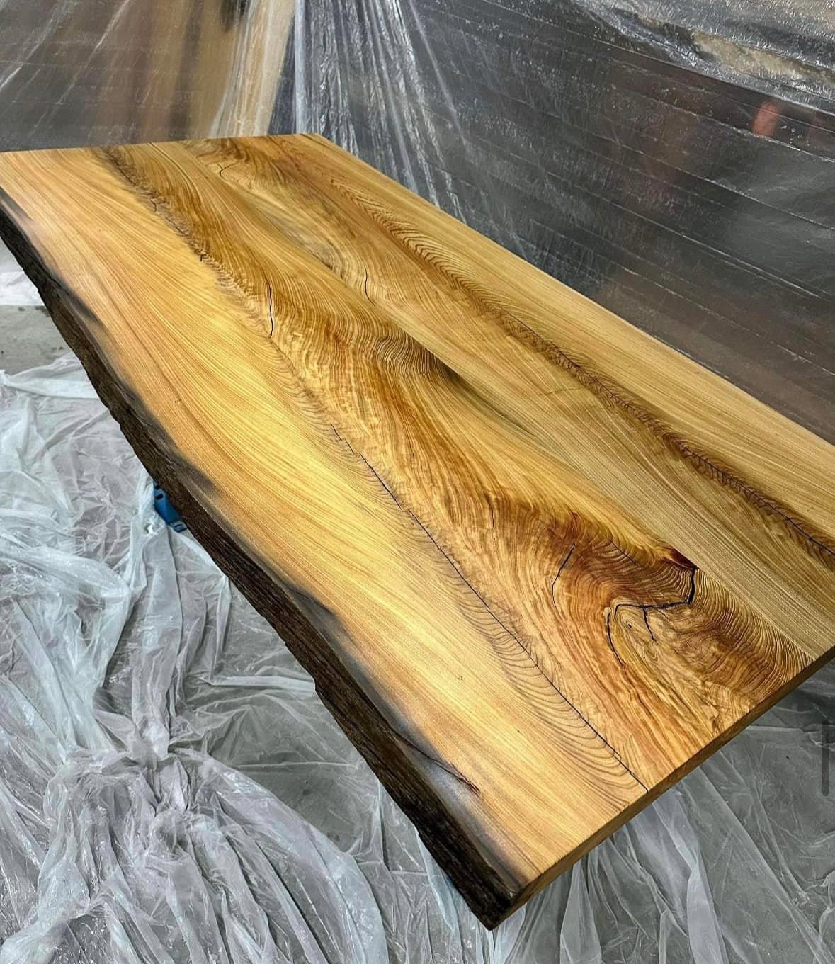 A polished live edge wooden slab with natural grain and cracks, resting on plastic sheeting.