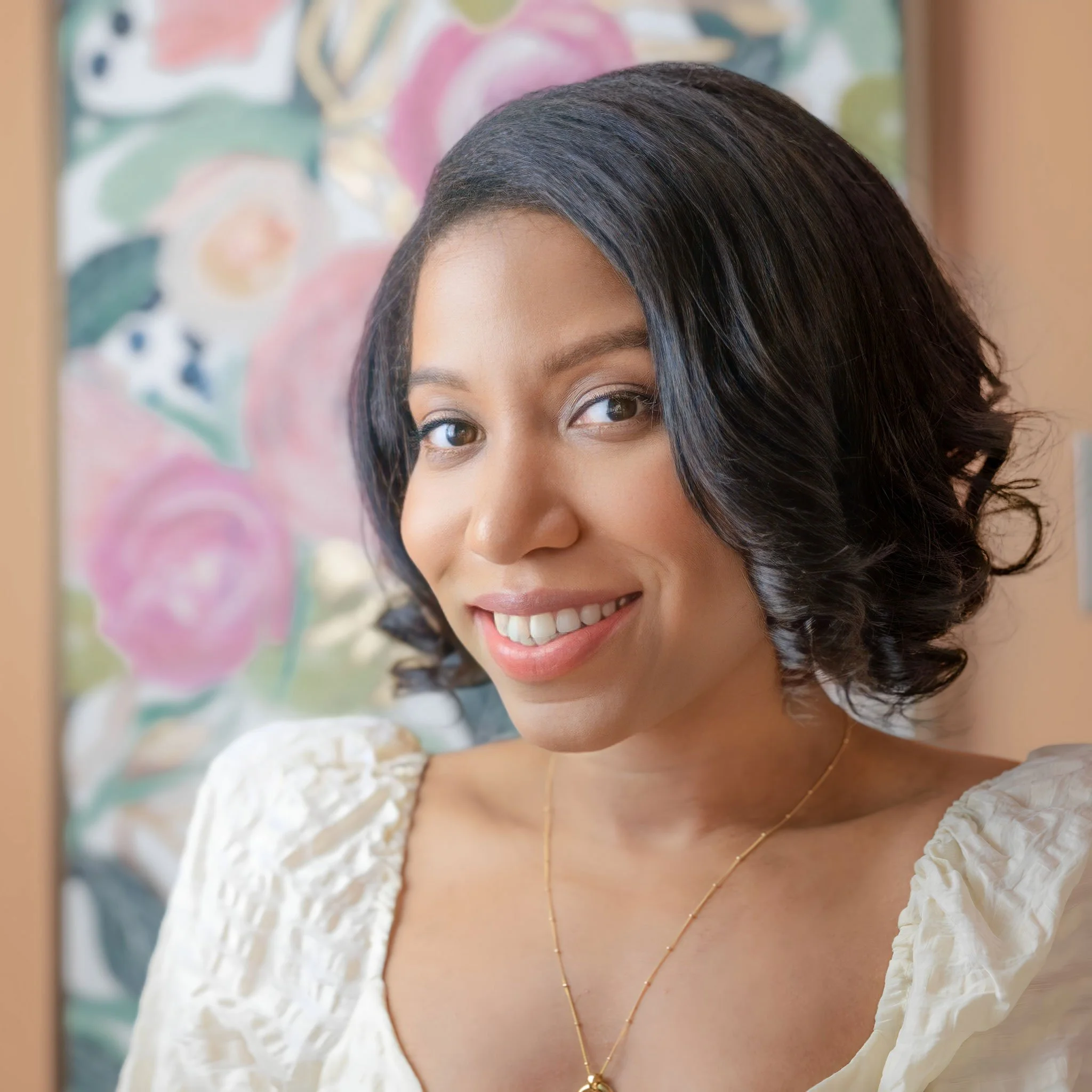 A woman with shoulder-length black hair smiling at the camera, wearing a white blouse and a gold necklace, with a colorful floral painting in the background.