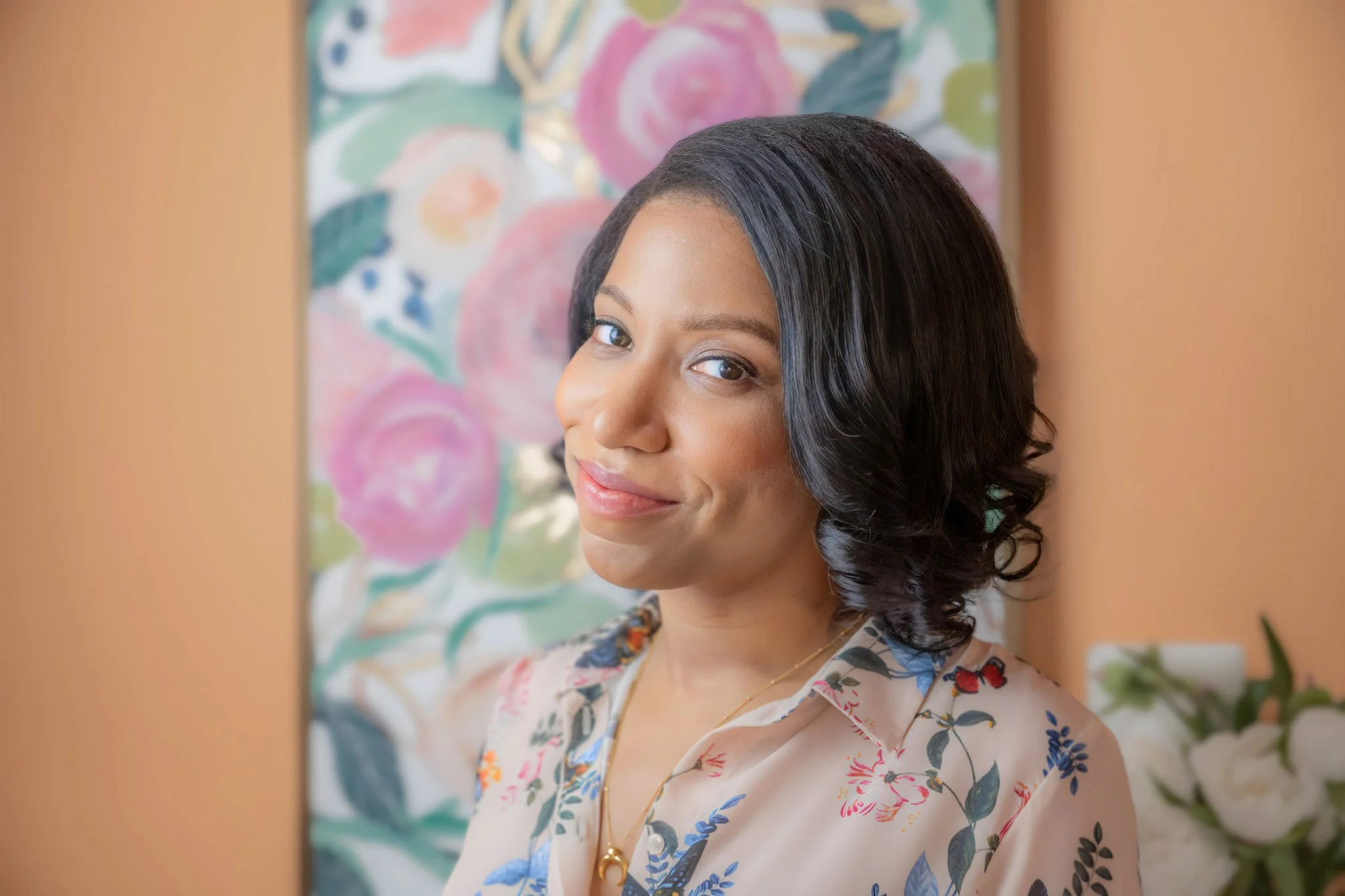 Gina Davis, a woman, with shoulder-length black wavy hair, wearing a floral blouse and jewelry, smiling slightly while looking at the camera. Background features a colorful floral painting.