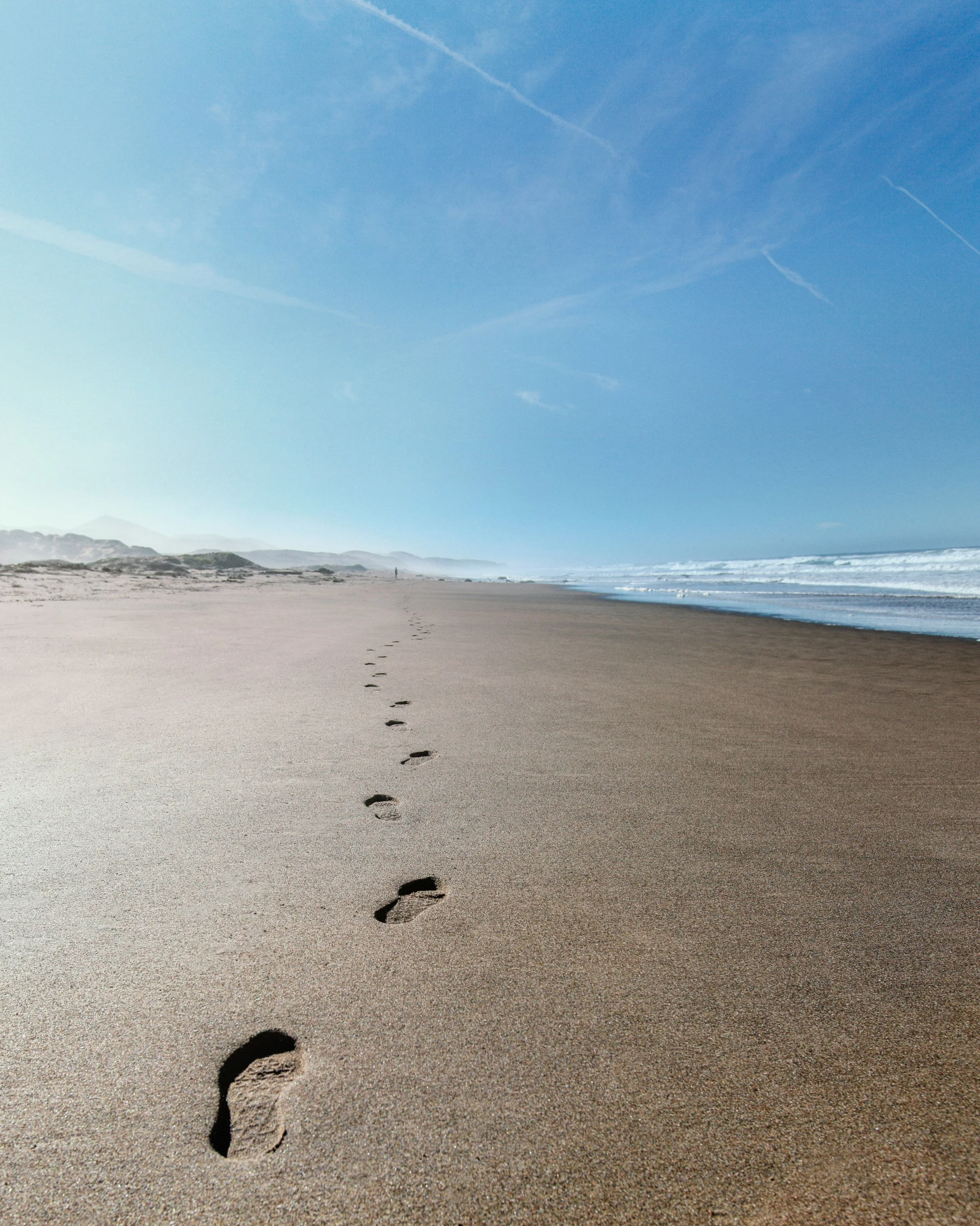 Footprints in the sand on a beach with the ocean on the right and a blue sky above.