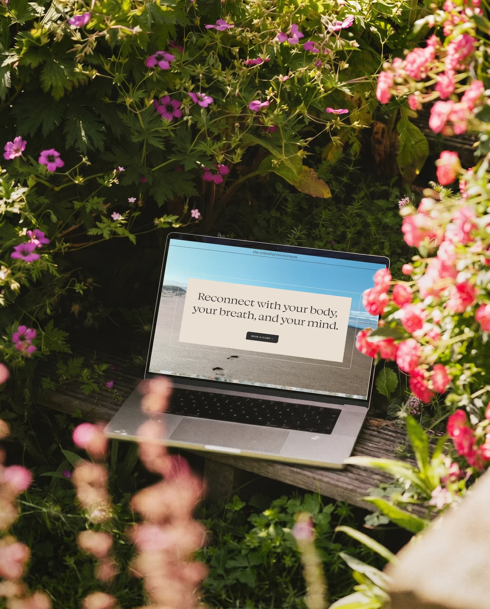 A laptop on a wooden bench surrounded by pink and purple flowers with a screen displaying an inspirational message about reconnecting with your body, breath, and mind.