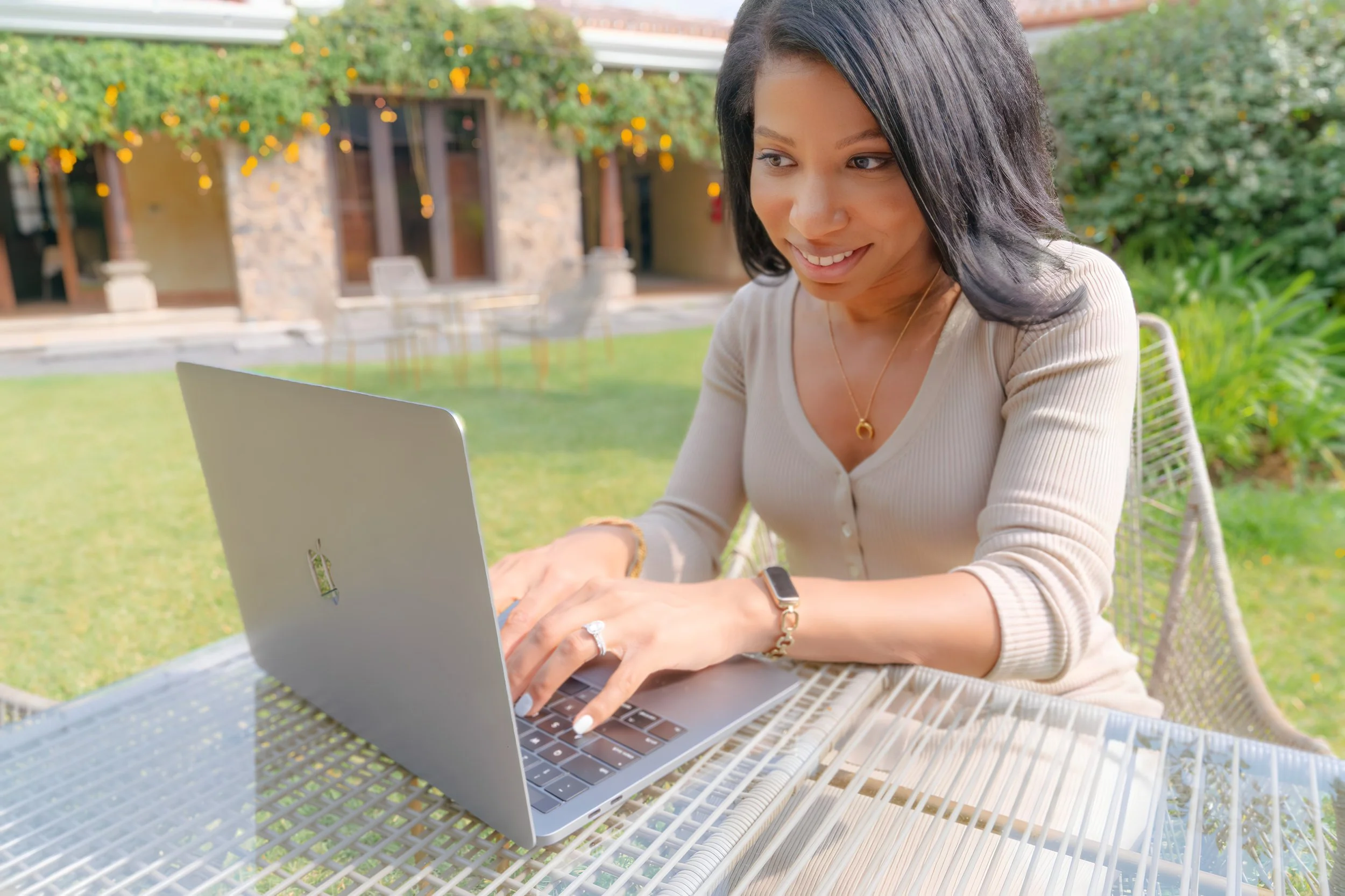 A woman, Gina S. Davis, sitting at a glass outdoor table with a silver laptop, smiling and typing, with a garden and house in the background.