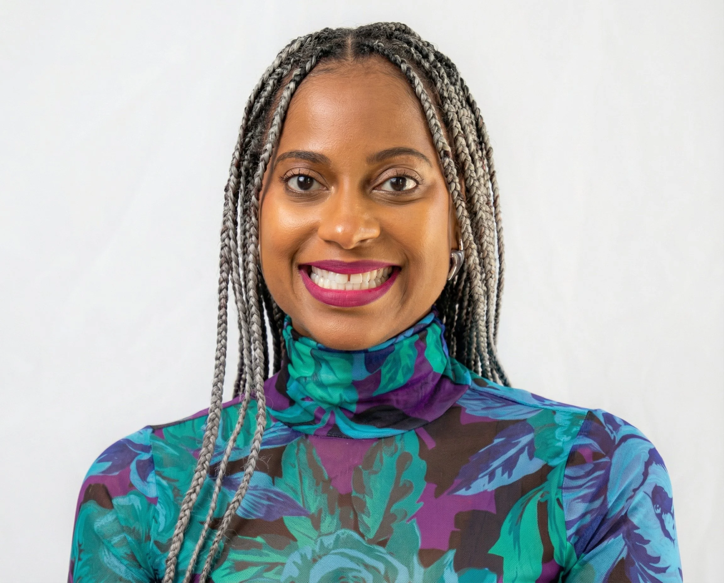 Close-up portrait of a woman with braids smiling, wearing a colorful, patterned high-neck top against a plain white background.
