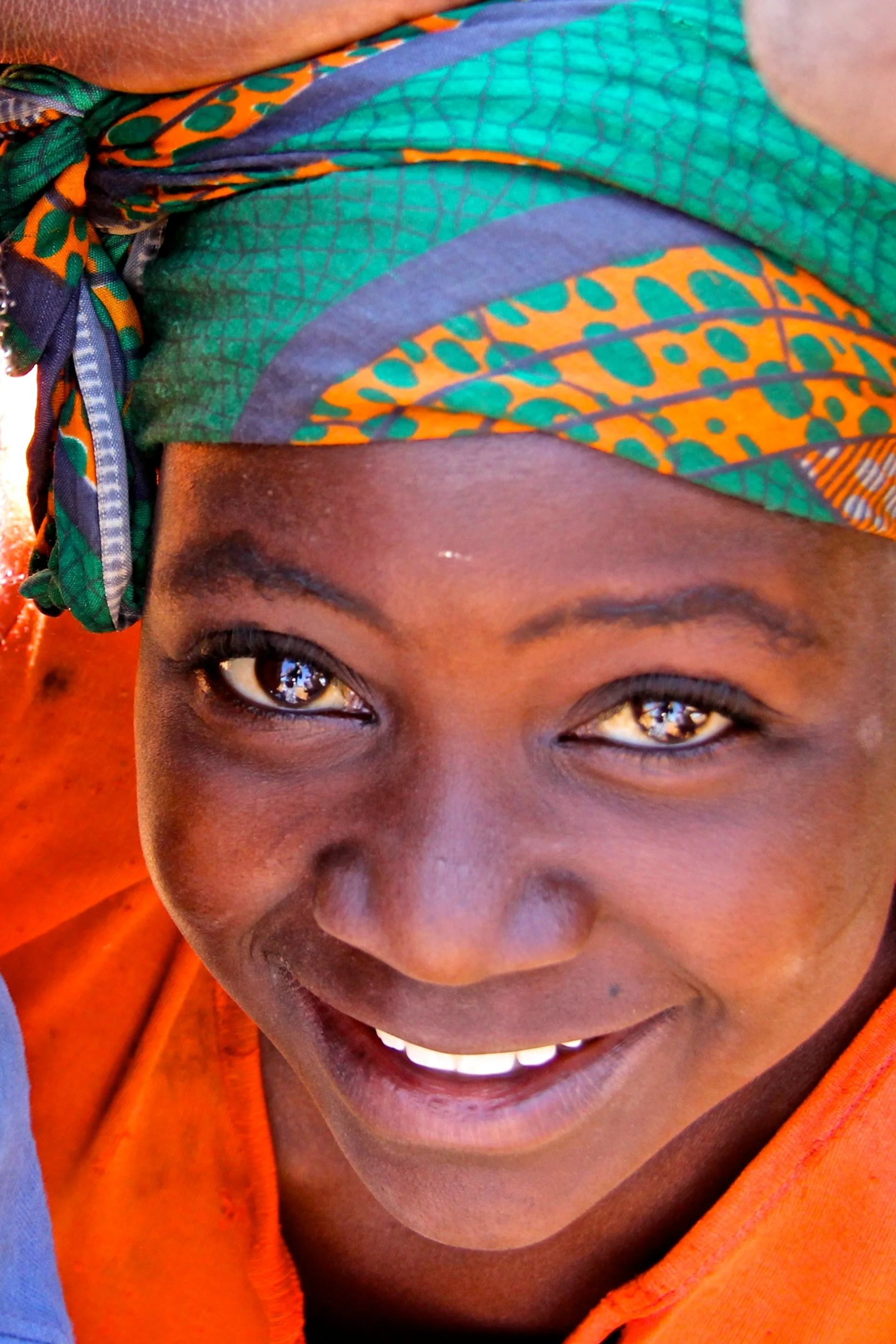 Close-up of a smiling person's face wearing a colorful headscarf and an orange shirt, with visible eyes and teeth.