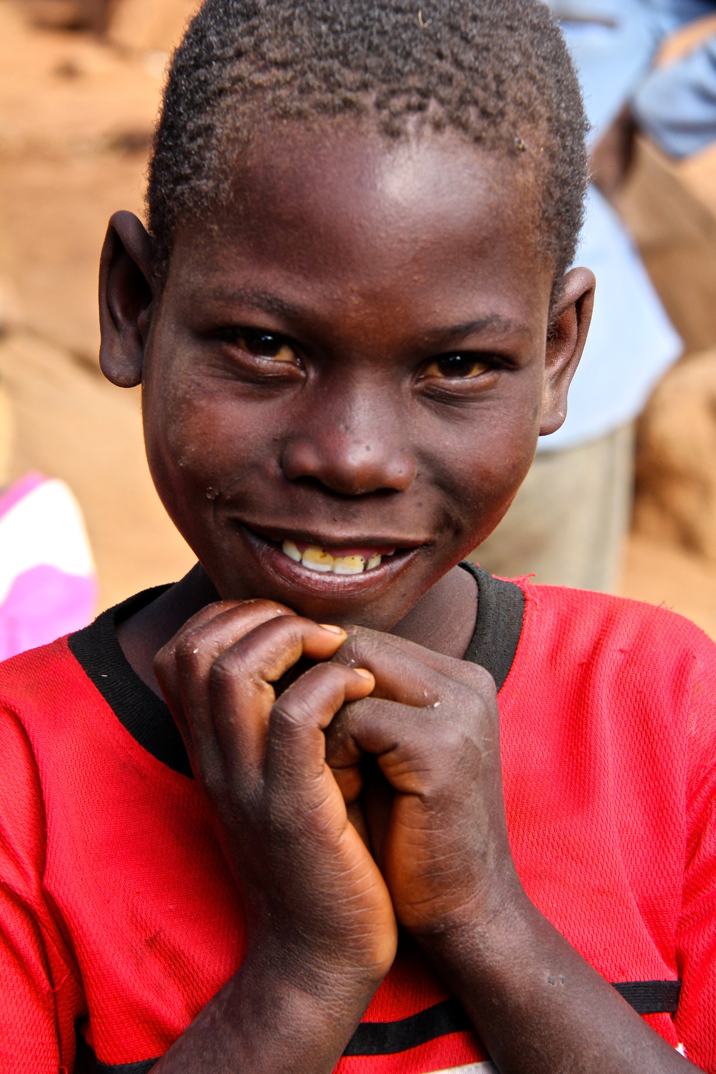 A close-up of a young boy with short curly hair, wearing a red shirt with black trim, smiling and holding his hands together near his chin, with a blurred background.