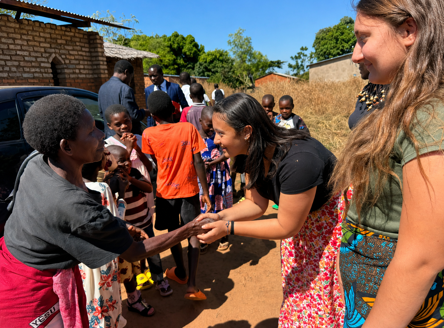 Two women, one black and one white, shaking hands and smiling in an outdoor rural setting with children and other adults around. The black woman is wearing a black shirt and a red skirt, while the white woman has long blonde hair and is wearing a khaki shirt and colorful patterned skirt. There is a brick building and green trees in the background.
