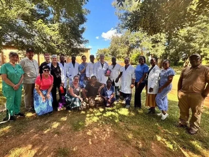 Group of healthcare professionals and staff standing outdoors on a sunny day, some in medical scrubs and others in casual or professional attire.