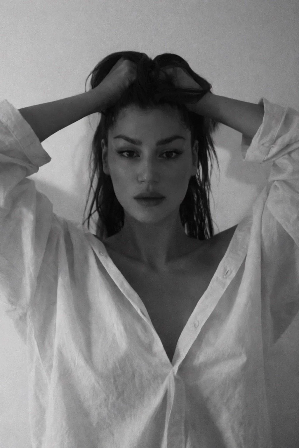 A woman with long, wet hair wearing a loose white button-up shirt, holding her head with both hands, looking directly at the camera, in black and white.