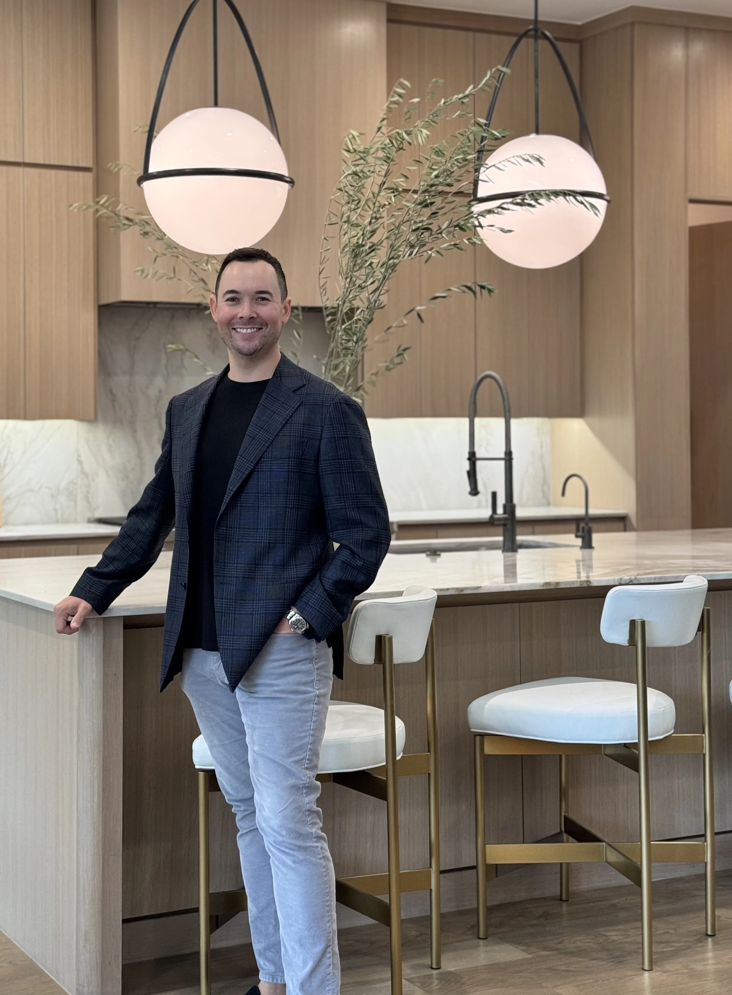 A man standing in a modern kitchen with a marble island, wooden cabinets, and hanging spherical pendant lights. He is smiling and wearing a dark blazer, black shirt, and light-colored pants.