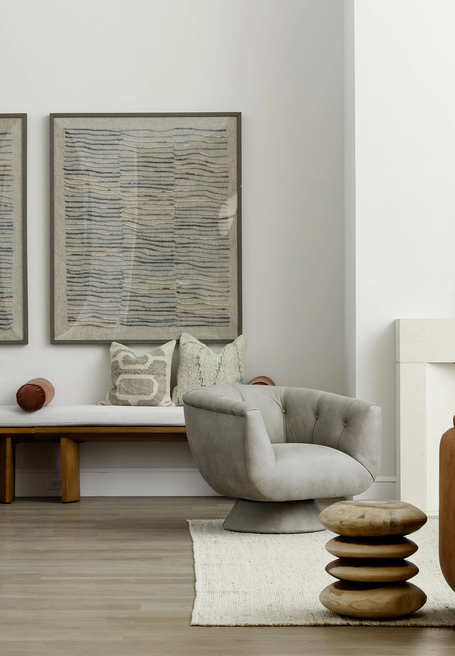 A modern living room corner with a beige armchair, a wooden side table with layered wooden design, a white bench with decorative pillows, and framed textured abstract art on the wall.