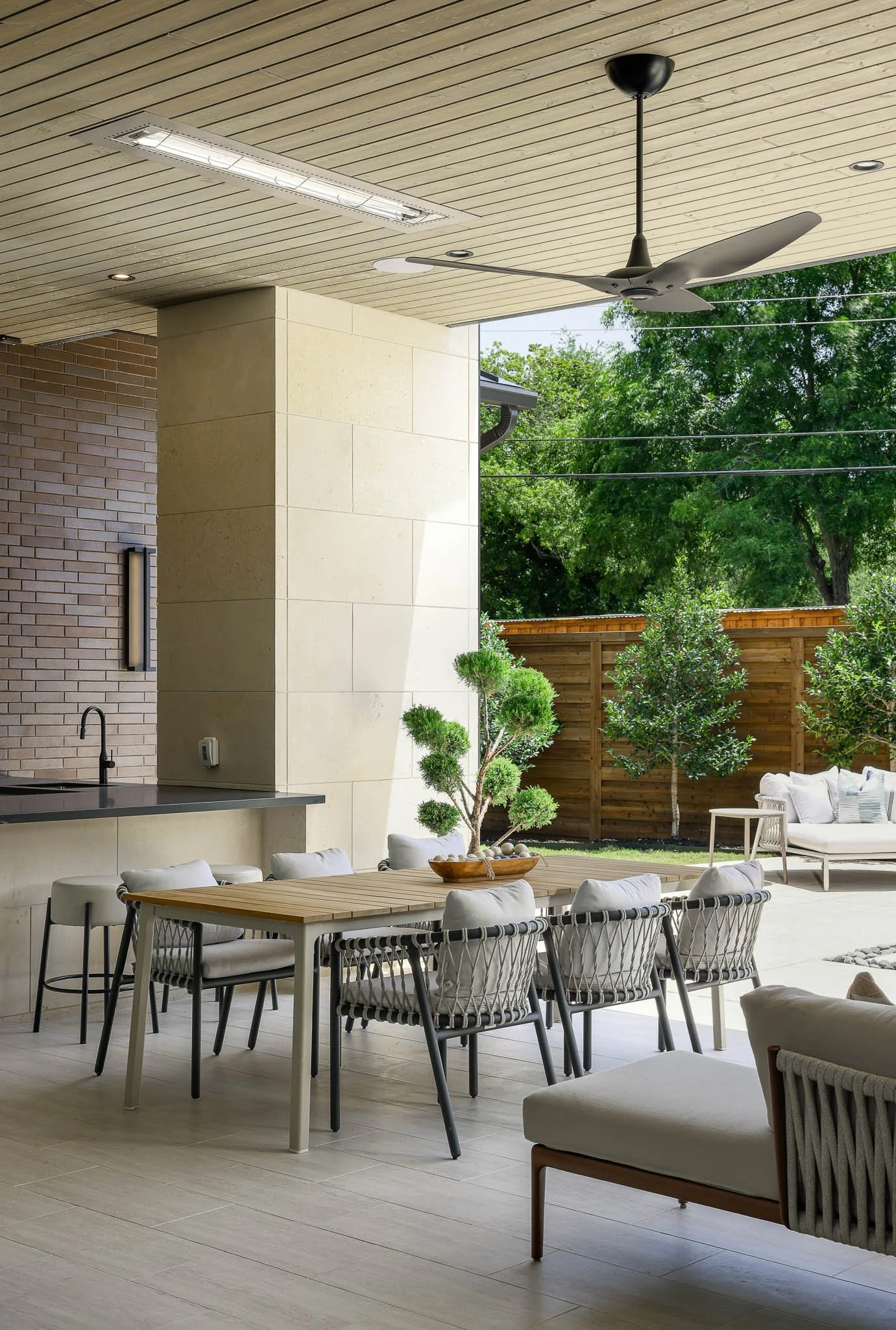 Modern outdoor patio with wooden dining table, cushioned chairs, and lounge area, surrounded by trees and a wooden fence.
