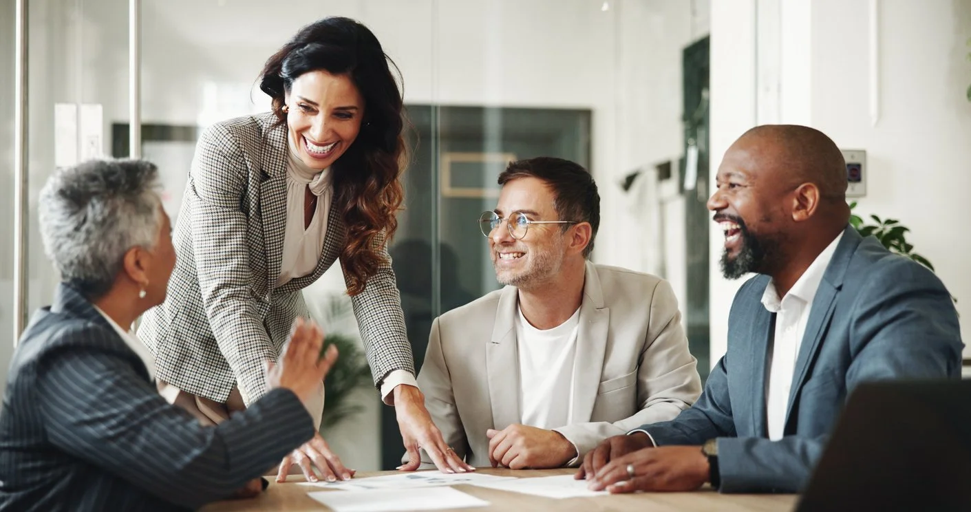 A diverse group of five professionals in a meeting room, smiling and engaging in conversation around a table.