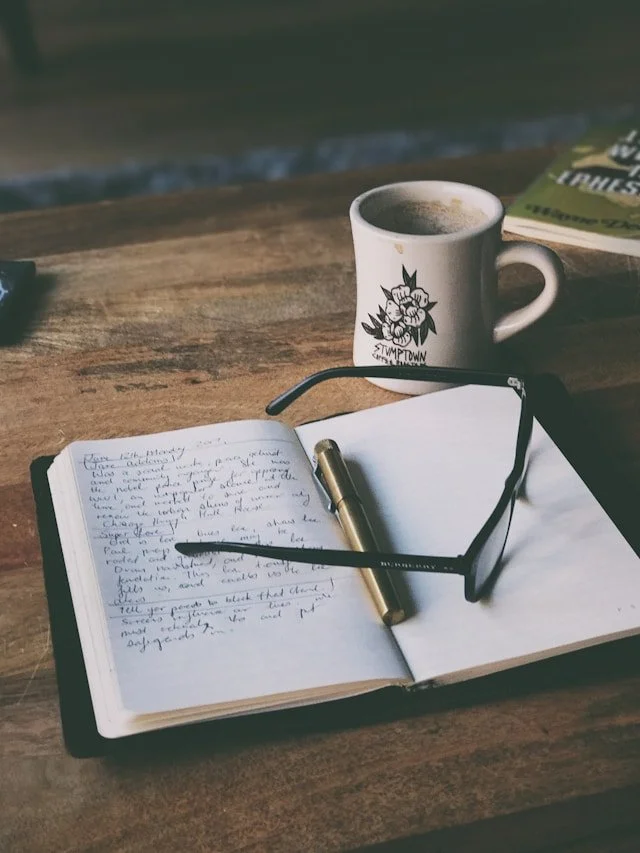 A cup of coffee on a rustic wooden table with a journal, a pen, and eyeglasses, and a magazine in the background.