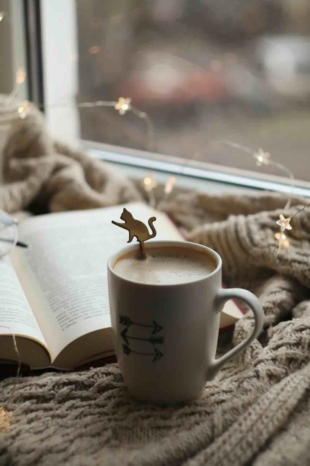 A warm mug resting on a thick knitted blanket next to an open book and glowing star-shaped string lights by a window, evoking a sense of deep comfort and a regulated nervous system.