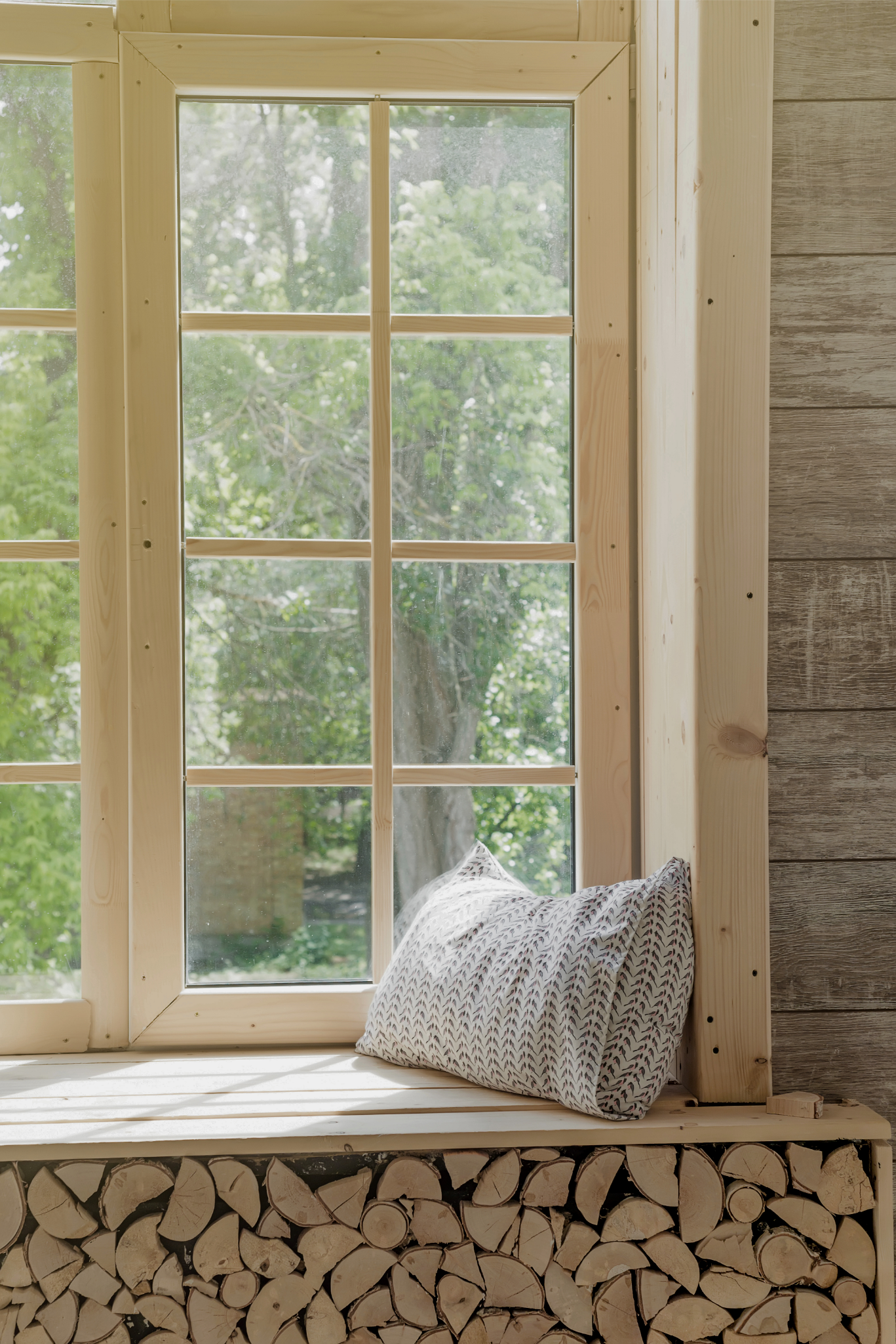 A wooden-framed window with a patterned pillow on the windowsill, overlooking green trees outside.