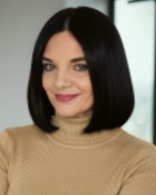 A woman with shoulder-length black hair wearing a beige turtleneck sweater, smiling, in an indoor setting.
