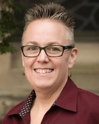 A woman with short, spiked gray hair, glasses, and earrings, smiling outdoors in front of a blurred background.