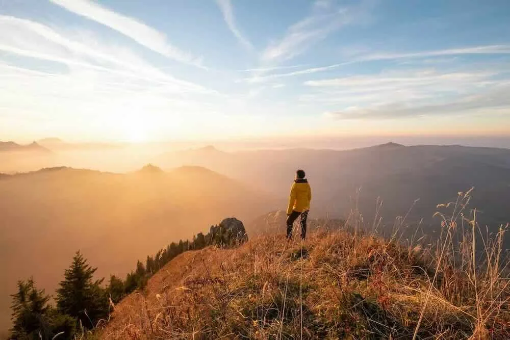 A person in a yellow jacket standing on a grassy mountain ridge overlooking a vast, foggy valley at sunrise, symbolizing clarity and overcoming emotional overwhelm and anxiety through therapy.