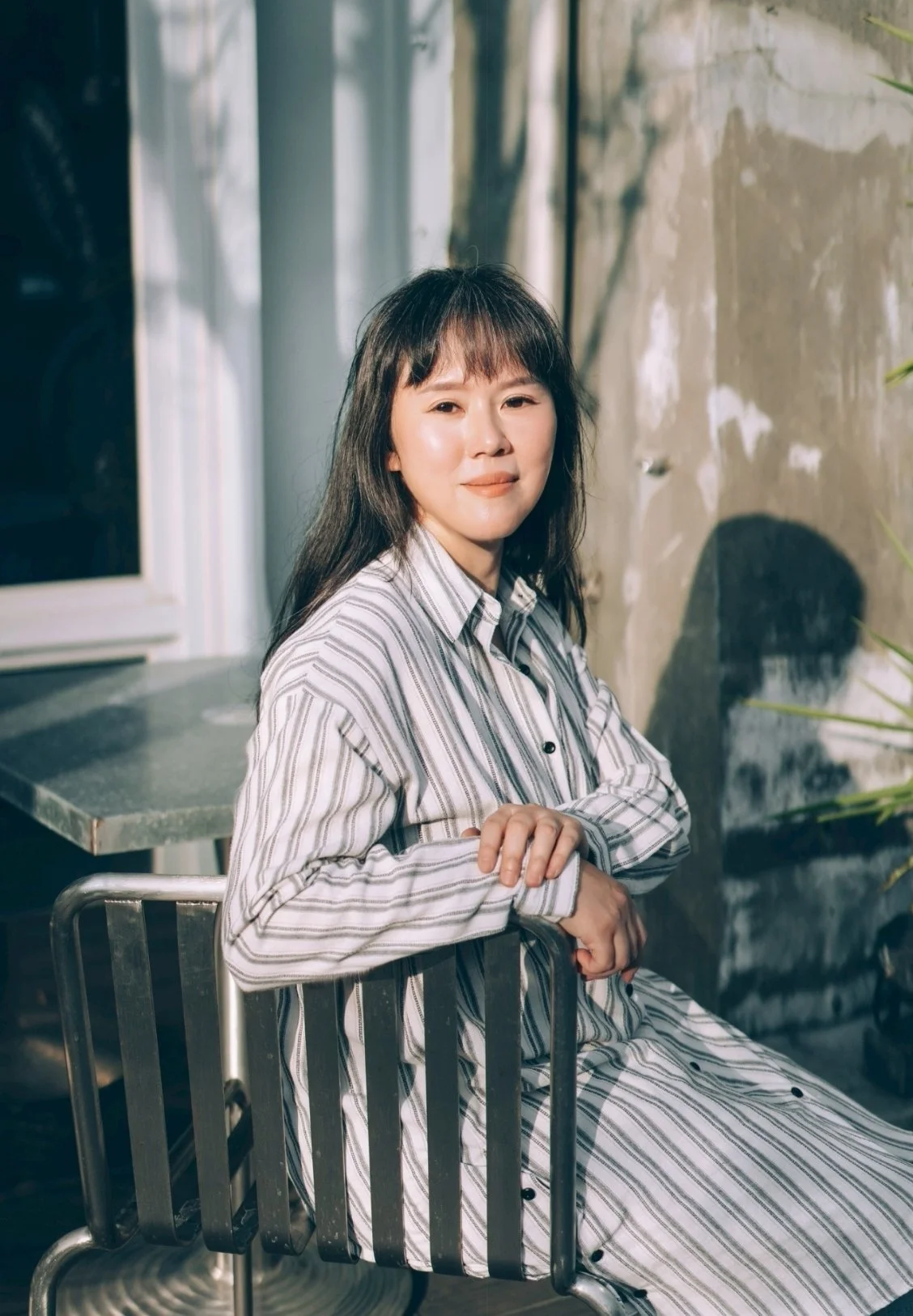 A woman with long dark hair wearing a striped shirt sitting on a metal chair near a wall with sunlight coming through a window.