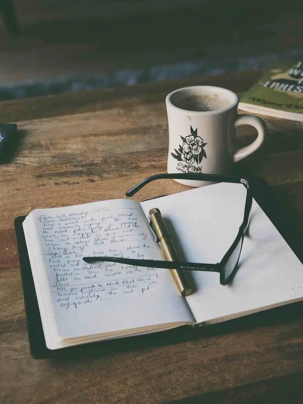An open journal with a pen and eyeglasses on a rustic wooden table next to a coffee cup, symbolizing the deep reflection and mental clarity achieved during an EMDR intensive.
