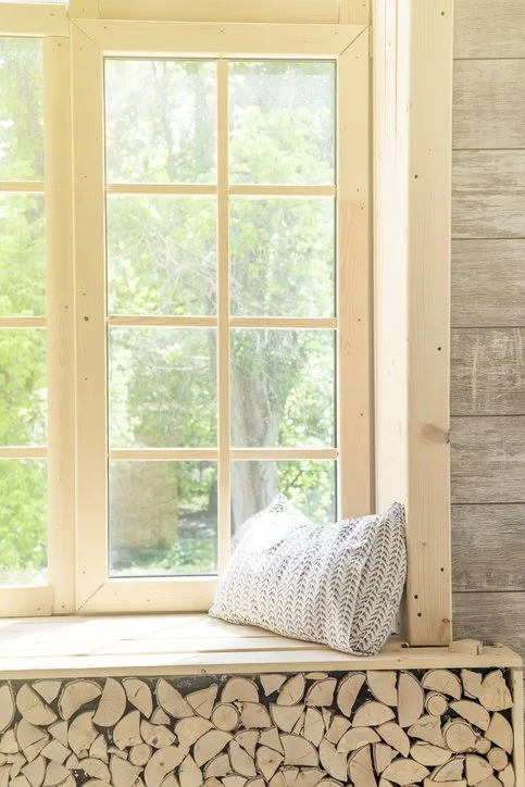 A cozy window nook with a geometric-patterned pillow, surrounded by a wood-paneled wall and a wood storage area filled with chopped firewood, overlooking a green outdoor scene.
