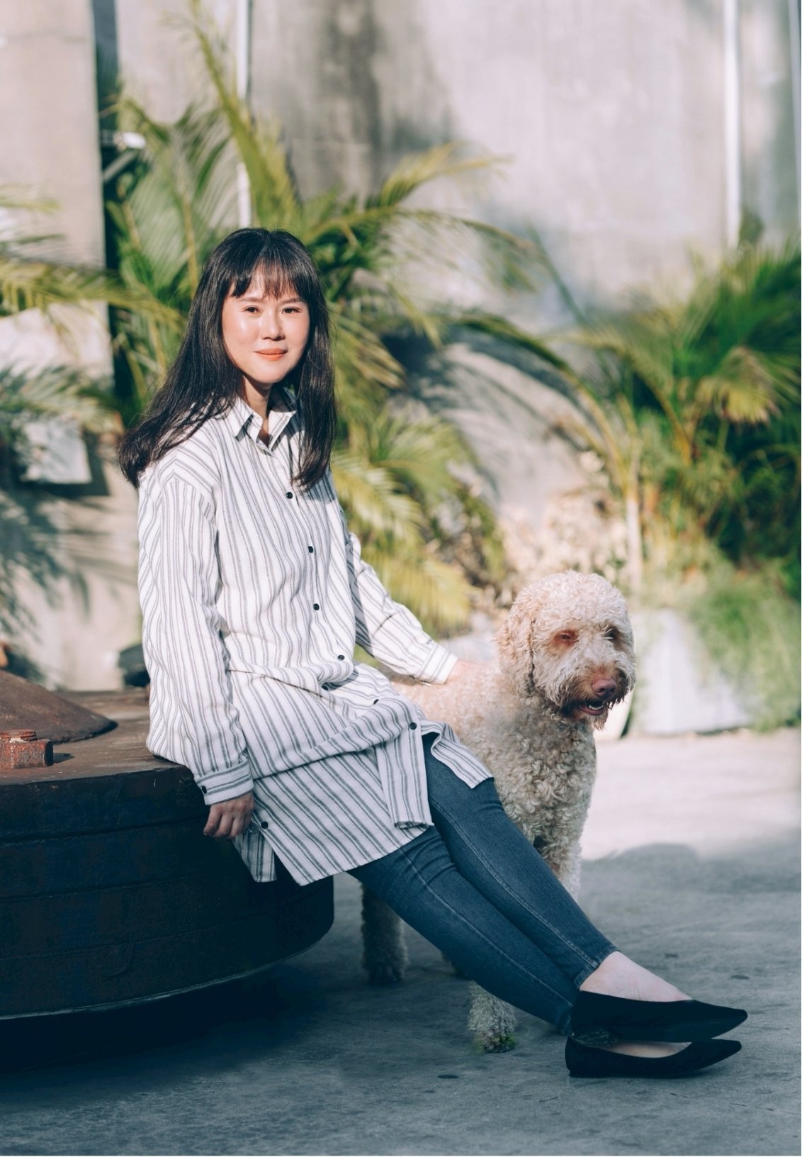 A young woman with dark hair and bangs, wearing a white and gray striped shirt and jeans, sitting on a tire while smiling. She has a large, curly-haired dog sitting beside her. They are outdoors with tropical plants and a concrete wall in the background.