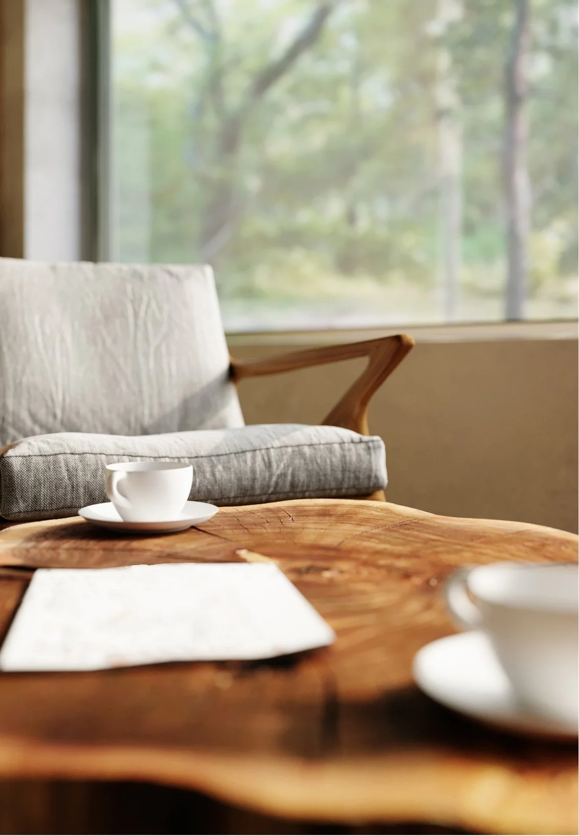 Empty white coffee cups on a wooden table in a cozy room with a sofa and large window showing trees outside.
