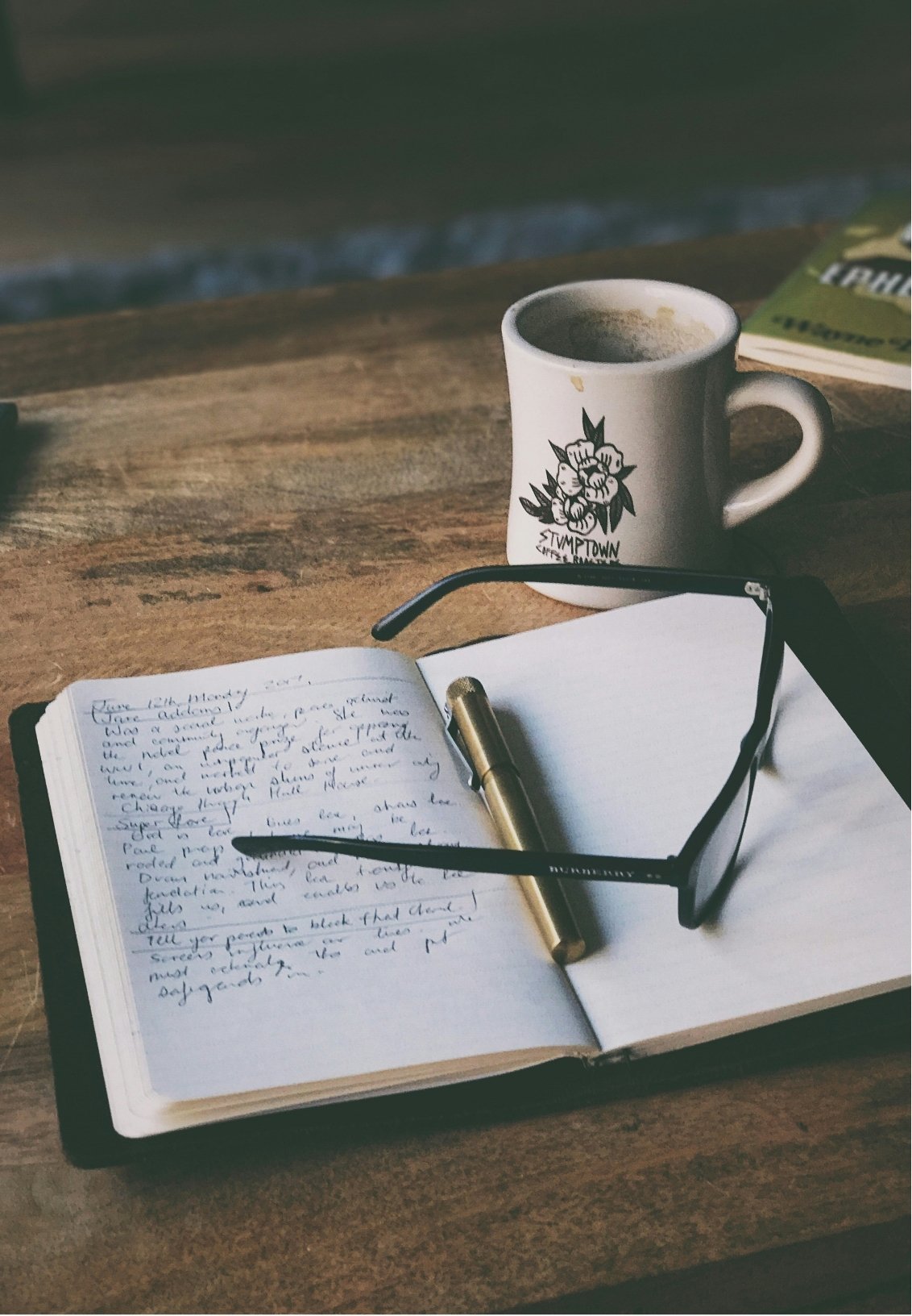 Open notebook with handwritten notes, black glasses, gold and black pen on a wooden table. Behind, a white mug with flowers and the words 'Stumptown Coffee Roasters' and a second, partially visible magazine or book.
