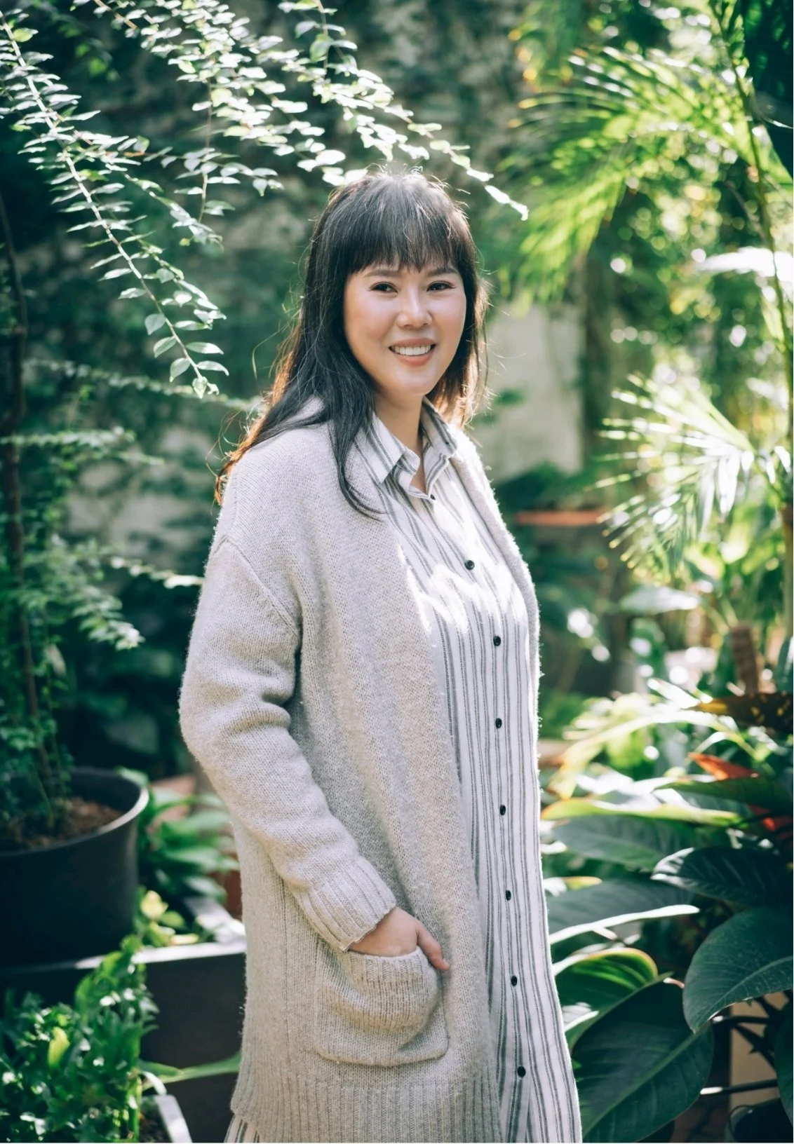 A smiling woman standing outdoors among lush green plants, wearing a light cardigan and a striped dress with her hands in her pockets.
