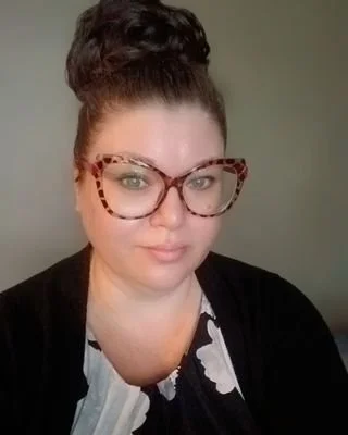 A woman with dark hair in a bun, wearing large tortoiseshell glasses and a black and white patterned top.