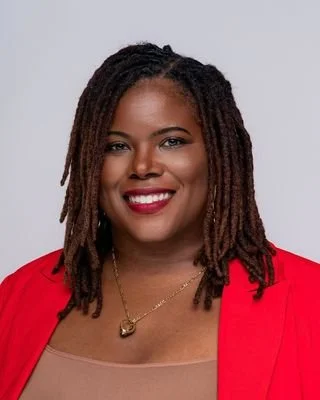 Portrait of a smiling woman with shoulder-length dreadlocks wearing a red blazer and gold necklace against a gray background.