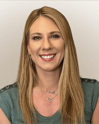 A woman with long blonde hair smiling and wearing a green top and layered necklaces.