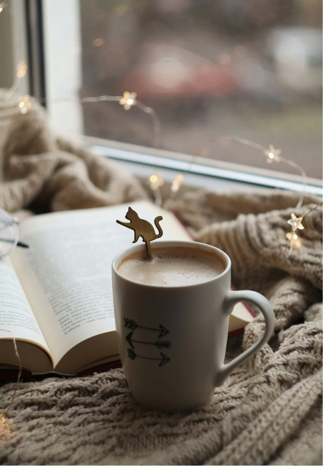 A cozy scene with a mug of hot beverage with a cat-shaped stirrer, an open book, a soft knitted blanket, and a string of star-shaped fairy lights by a window.