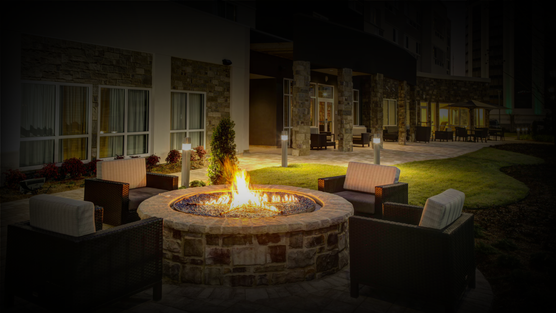 Outdoor patio at night with a round fire pit table and surrounding chairs, exterior of a building with large windows, landscaped greenery, and string lights.