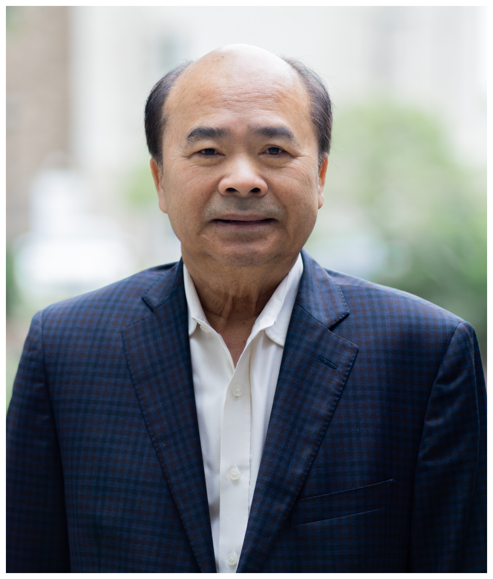 A middle-aged Asian man wearing a dark blue checkered blazer and white shirt, standing outdoors with blurred greenery in the background.