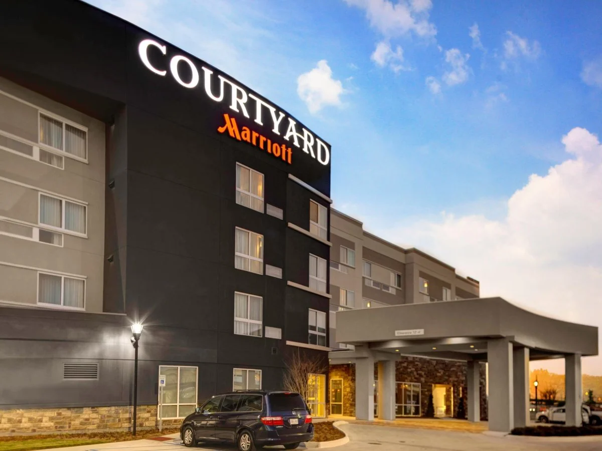 Exterior view of the Courtyard Marriott hotel showing the hotel sign, entrance, parked cars, and a sunny sky at dusk.