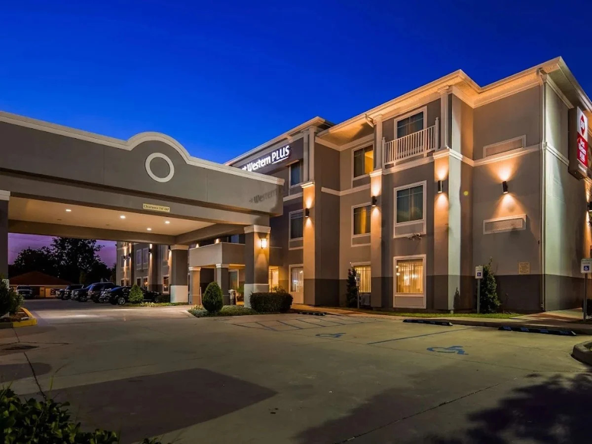 A modern hotel building at dusk with exterior lighting illuminating the walls and parking lot in the foreground.