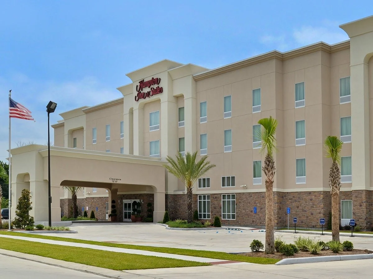 Exterior of a Hampton Inn & Suites hotel with palm trees, a parking lot, and an American flag, under a partly cloudy sky.