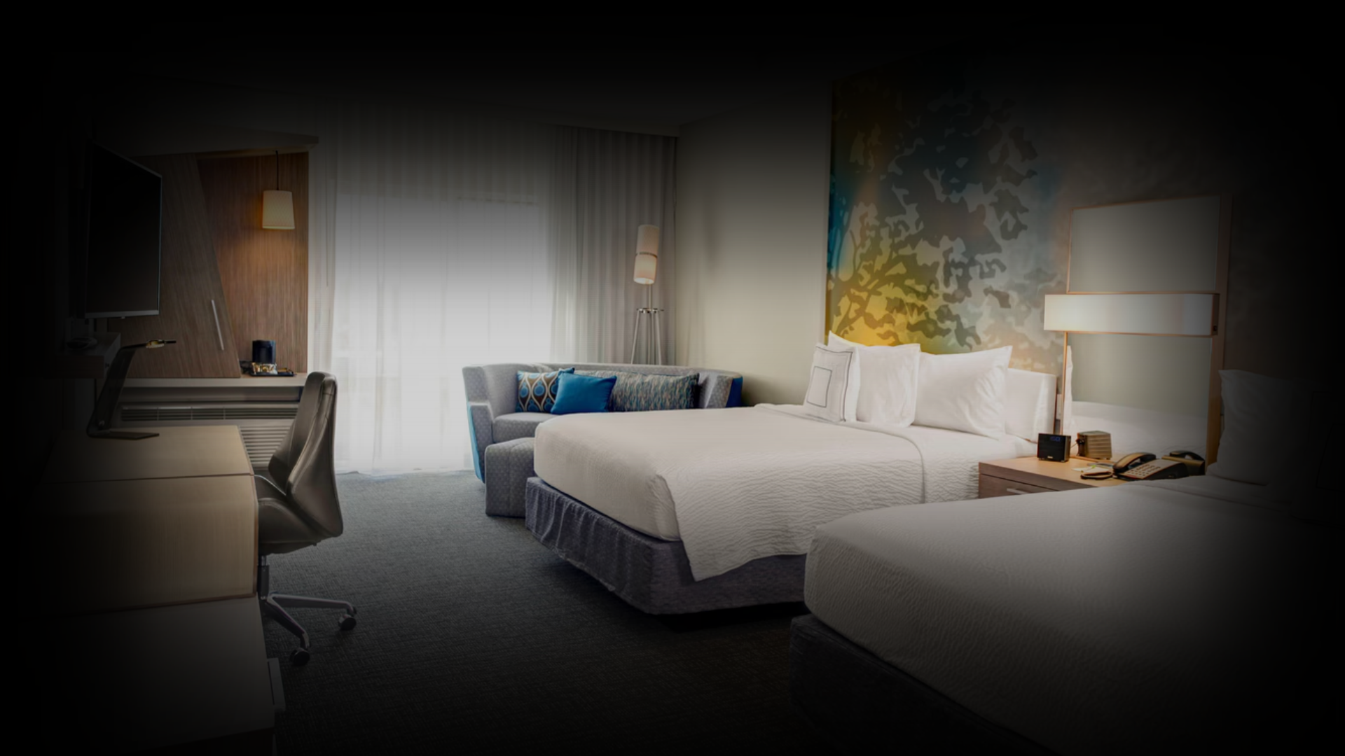Interior of a modern hotel room with two beds, a couch, a desk, and a window with curtains.