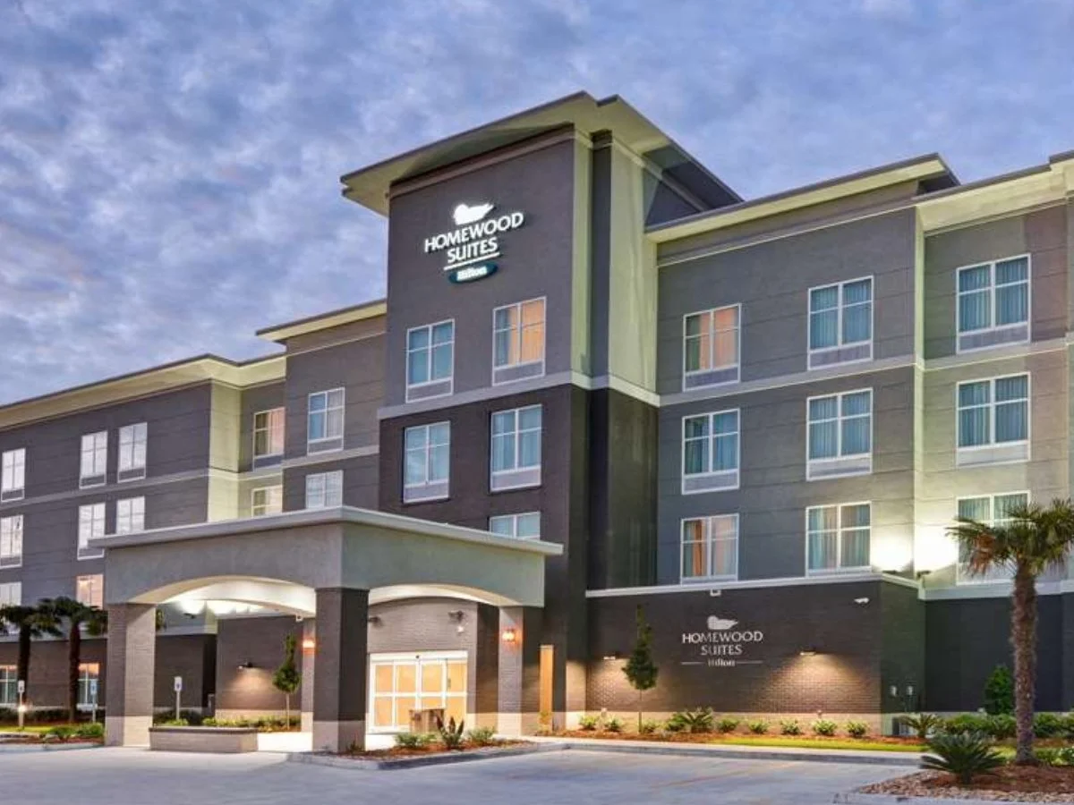 Exterior view of a modern four-story hotel building with a sign reading 'Homewood Suites by Hilton,' featuring a large entrance, landscaping, and palm trees.