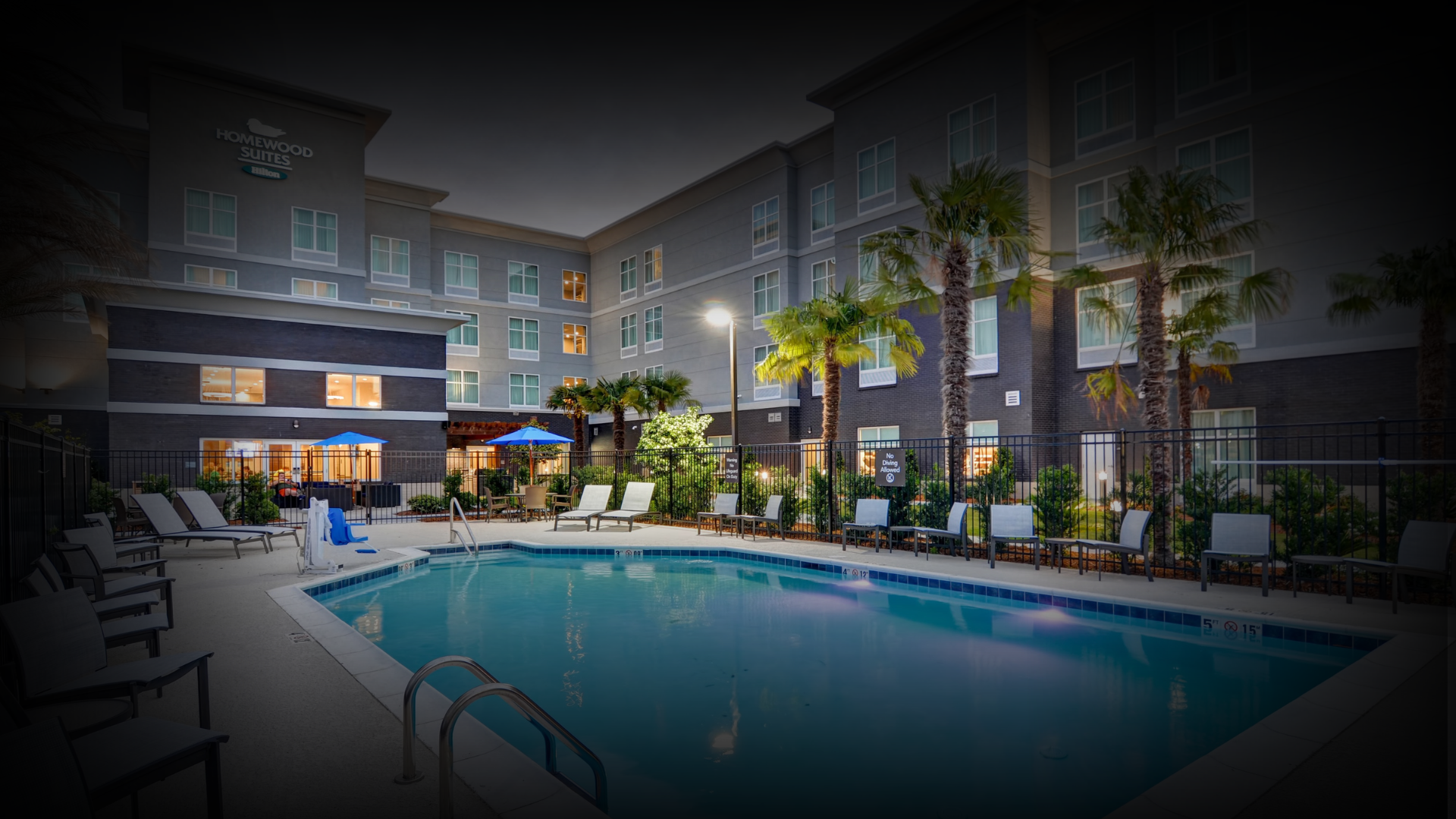 Nighttime view of a hotel pool area with lounge chairs, umbrellas, and surrounding palm trees, with a multi-story hotel building in the background.