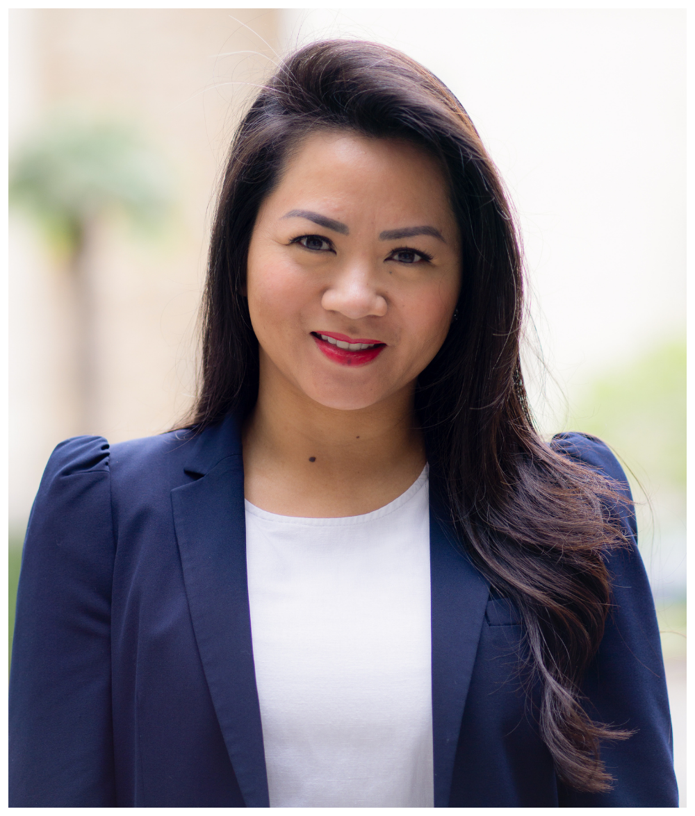 A woman with long dark hair wearing a navy blazer and a white top, smiling outdoors with blurred greenery in the background.