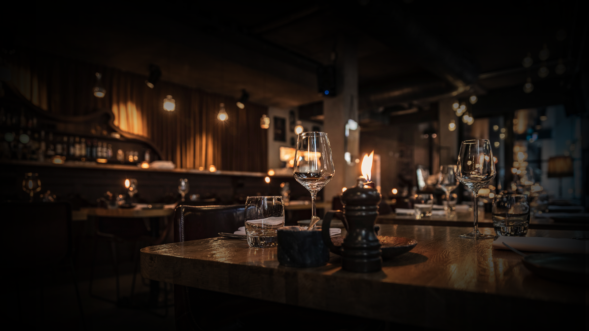 Dimly lit restaurant with wine glasses, water glasses, and candles on wooden tables, cozy atmosphere with warm lighting and dark decor.