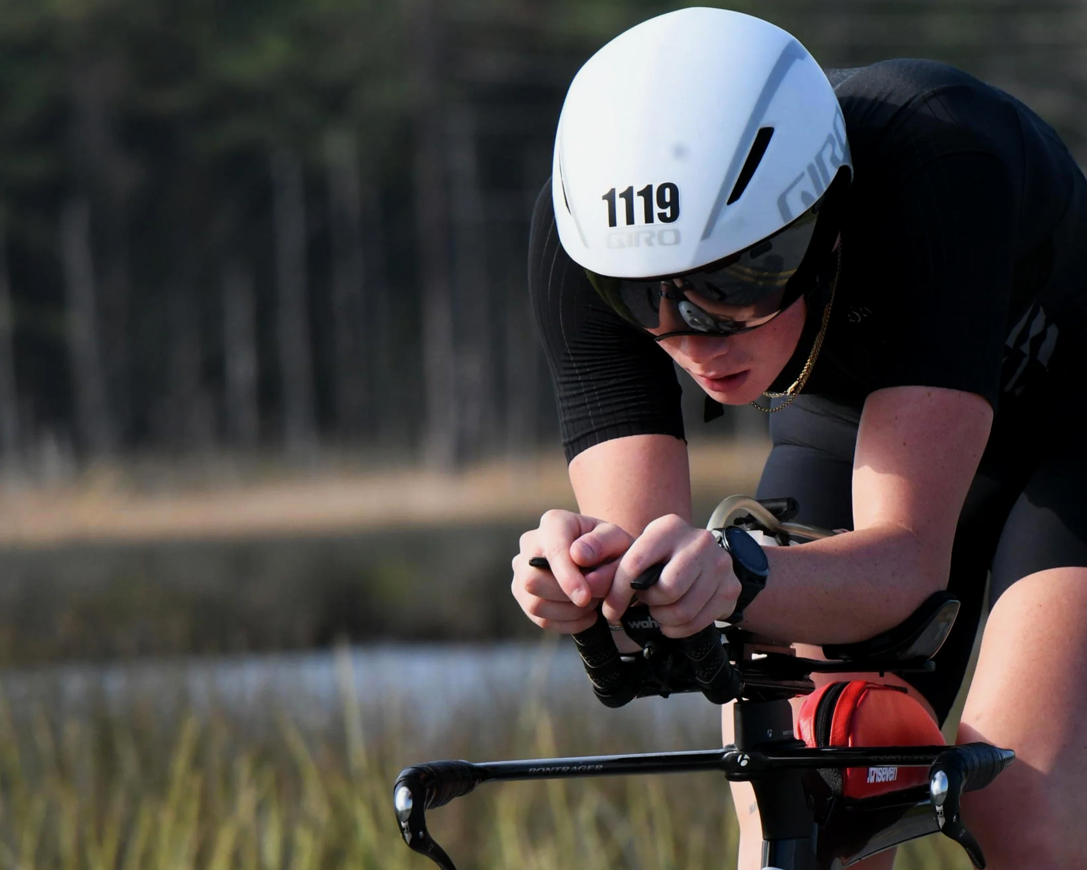 Close-up of a cyclist wearing a white helmet with the number 1119, black sunglasses, and a black cycling outfit, riding outdoors.