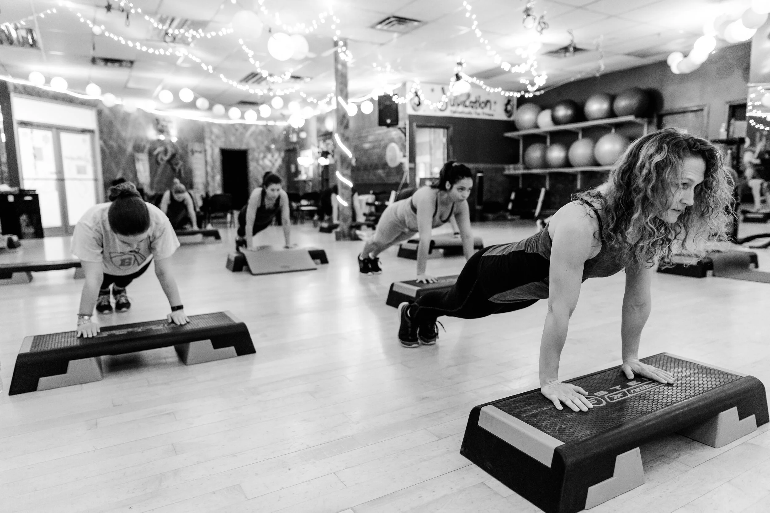 A group of five women participating in a fitness class doing push-up exercises on aerobic steps in a gym with string lights and fitness equipment in the background.