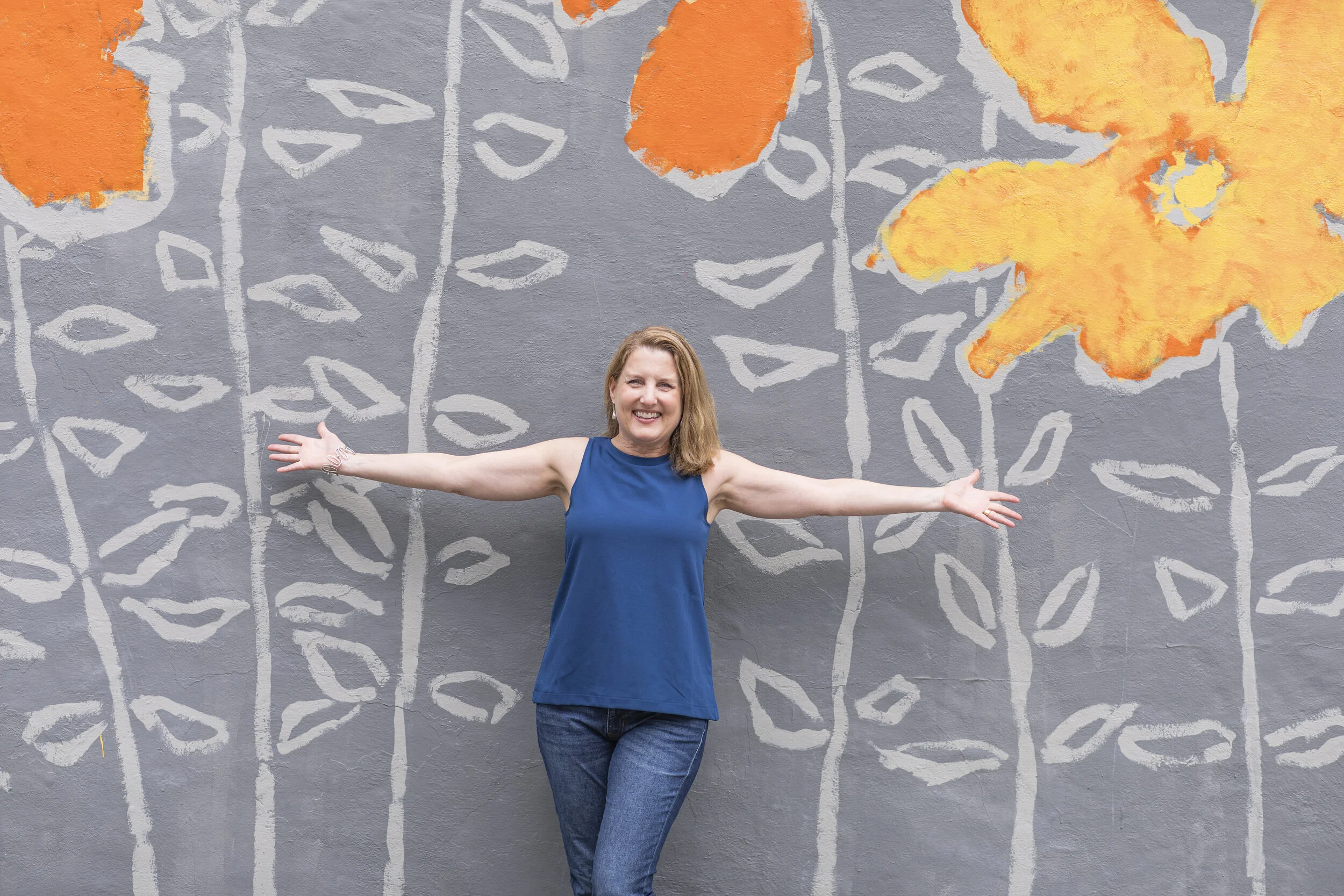 Woman in a blue sleeveless top and jeans stands in front of a gray wall with orange and yellow leaf paintings, smiling with arms outstretched.