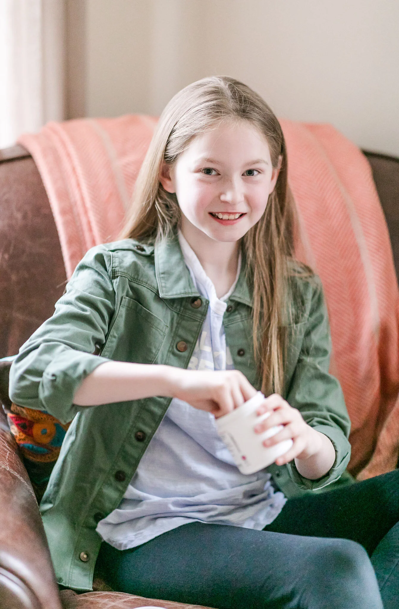 A young girl with long light brown hair, wearing a green jacket and a white shirt, smiling and sitting on a brown couch holding a white mug.