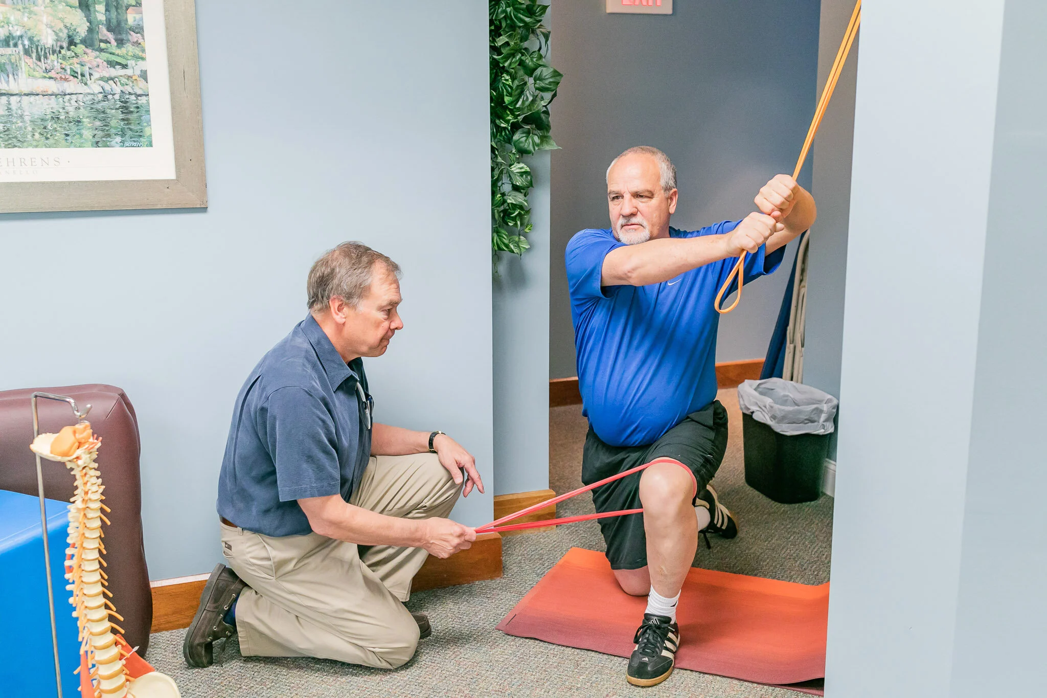 A man in a blue t-shirt demonstrating a knee stretch with resistance bands assisted by another man in a gray shirt, in an office or medical setting.