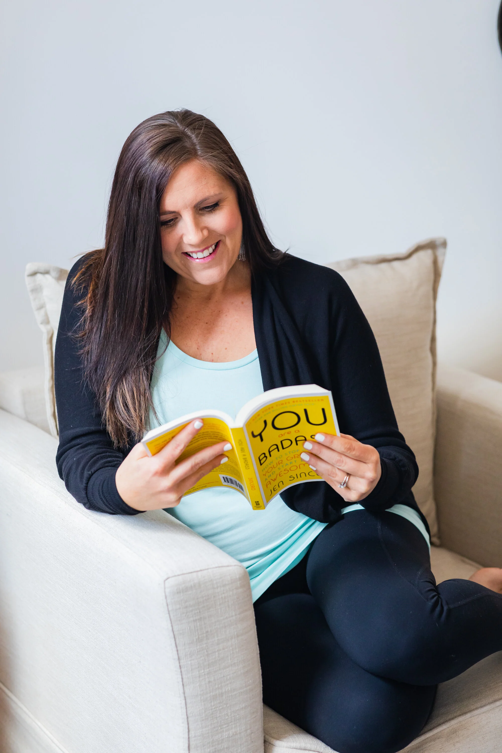 Woman sitting on a couch, smiling, and reading a yellow-covered book.