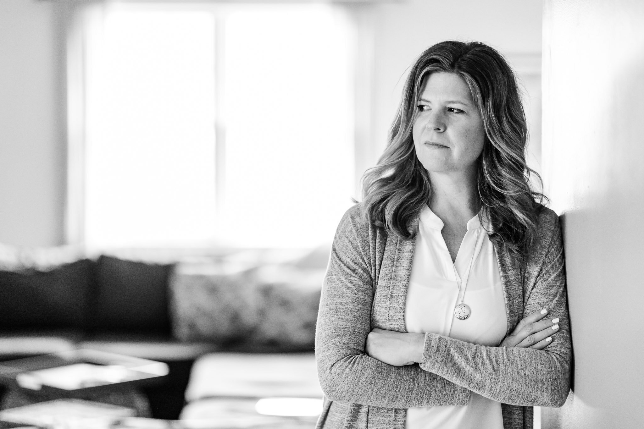 A woman with wavy hair looks to the side with a serious expression, standing against a wall in a bright room.